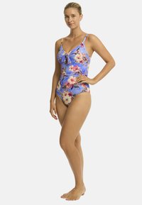 Een vrouw staat blootsvoets in een blauw badpak met bloemenpatroon, glimlachend met haar hand op haar heup.