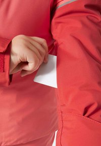 Giacca rossa realizzata in un tessuto liscio e resistente. Una mano sta inserendo una carta bianca in una tasca sul braccio, con dettagli riflettenti visibili.