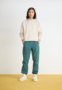 Beige hoodie met een voorkantzak, gecombineerd met teal tapered pants. Het schoeisel bestaat uit witte sportschoenen met accenten.
