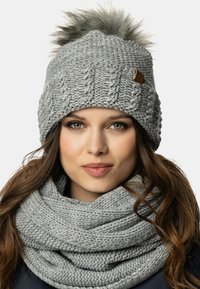 Cappello lavorato a maglia grigio con pom-pom di pelliccia e etichetta in pelle, abbinato a una sciarpa infinita grigia lavorata a maglia coordinata. Filato spesso e strutturato.