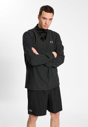 Veste de sport noire avec un col haut, devant zippé et logo ; associée à un short noir assorti. Texture lisse, design fonctionnel.