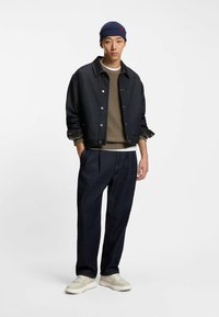 Dunkelblaue Jeansjacke mit silbernen Knöpfen, olivgrüner Strickpullover, marineblauer Beanie, gefaltete dunkelblaue Jeanshose und beiger Sneaker.