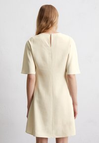 Kurzärmliges Kleid in knielanger Form in zartem Gelb. Verfügt über einen runden Halsausschnitt mit einer Schlüssellochöffnung im Rücken und einem glatten, strukturierten Stoff.