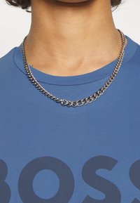 Silver kedja med växelvis släta och texturerade länkar, bärs över en blå crew-neck t-shirt med ett stort mörkt textmönster.