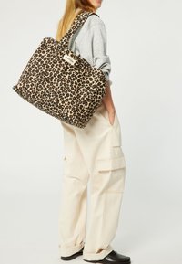 Sac fourre-tout en imprimé léopard avec un tissu texturé, doté d'une grande ouverture, de deux poignées robustes, et d'une étiquette de marque visible.