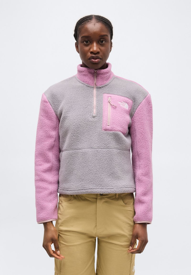 Donna che indossa un pile rosa e grigio con tasca sul petto con cerniera e pantaloni beige, rivolta in avanti su uno sfondo neutro.