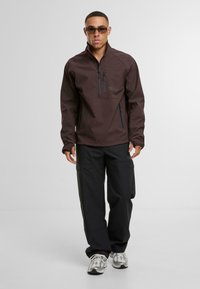 Braune Halbzip-Jacke mit Reißverschluss-Brusttasche, kombiniert mit schwarzen Cargo-Hosen. Das Modell trägt außerdem Sonnenbrillen und gemusterte Sneakers. Neutrale Hintergrund.