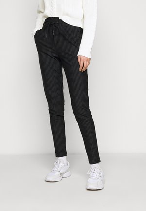 Persoon die zwarte broek met krijtstreepjes, witte gebreide trui, witte sokken en witte sneakers draagt, staand tegen een eenvoudige lichte achtergrond.