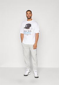 Camiseta blanca con un gráfico de los New York Yankees, combinada con pantalones cargo gris claro y zapatillas blancas con detalles en negro.