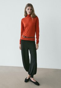 Frau trägt einen roten Rollkragenpullover, schwarze lockere Hosen mit Bündchen, einen schmalen braunen Gürtel und schwarze flache Schuhe, steht auf einem neutralen Boden.