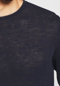 Detailní záběr na osobu, která má na sobě tmavě modrý pletený svetr s kulatým výstřihem a texturovanou látkou.