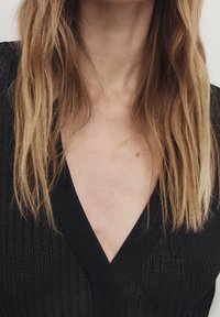 Schwarzes geripptes Strickoberteil mit tiefem V-Ausschnitt. Lange, wellige Haare, die teilweise sichtbar sind und die Textur und das Design des Kleidungsstücks betonen.