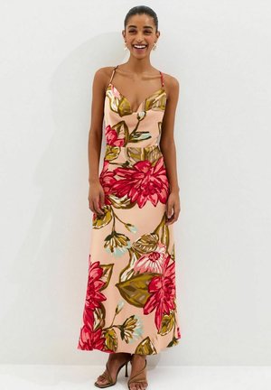 Woman smiling, wearing a sleeveless, peach-colored maxi dress with large red and green floral print and strappy brown high heels.