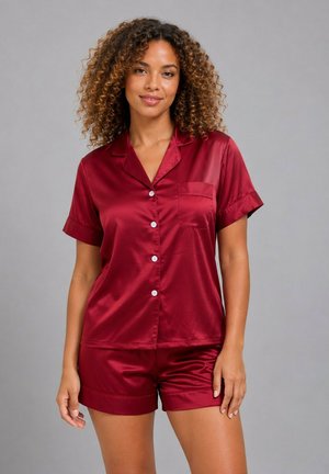 Femme aux cheveux bouclés portant un pyjama rouge en satin à manches courtes, boutonné, avec une poche poitrine, debout devant un fond gris uni.