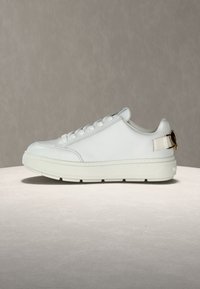 Λευκό χαμηλού ύψους αθλητικό παπούτσι με χοντρή σόλα, κορδόνια στο μπροστινό μέρος και λεπτομέρεια με χρυσή αγκράφα στο λουράκι της φτέρνας, εκτεθειμένο σε ουδέτερη επιφάνεια.