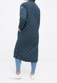 Femme portant un long manteau matelassé bleu foncé avec des boutons latéraux, un jean bleu clair et des baskets blanches, debout les mains dans les poches.