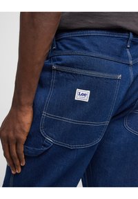 Blue denim jeans with a visible Lee label on the back pocket, contrasting white stitching, and a relaxed fit. Textured fabric.
