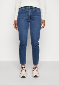 Blue denim jeans with a slim fit, high waist, reinforced pockets, and minimal fading, paired with light-colored sneakers.
