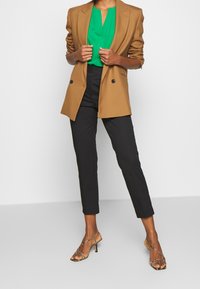 Brązowa dopasowana marynarka, zielona bluzka, czarne dopasowane spodnie oraz sandały na wysokim obcasie z paskami w wzór zwierzęcy.