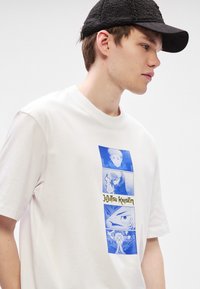 Λευκό βαμβακερό T-shirt με μπλε γραφικό σχέδιο που περιέχει τέσσερις πίνακες από το "Jujutsu Kaisen" και κίτρινο κείμενο. Στρογγυλή λαιμόκοψη, κοντά μανίκια.