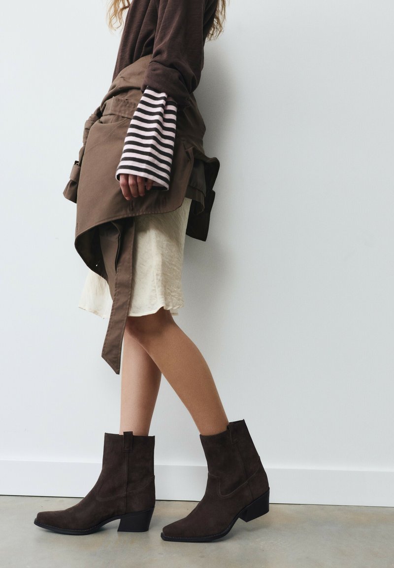 Femme portant des bottines marron, une jupe crème, une veste marron nouée autour de la taille et un haut rayé à manches longues, debout contre un mur blanc.