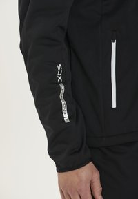 Sort softshell jakke med slim fit, glat tekstur, hvide lynlåse og "ENDURANCE XCS" mærkning på venstre ærme.