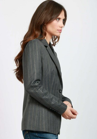 Donna con lunghi capelli castani, che indossa un blazer grigio a righe sottili e jeans blu, in piedi di profilo contro uno sfondo semplice.
