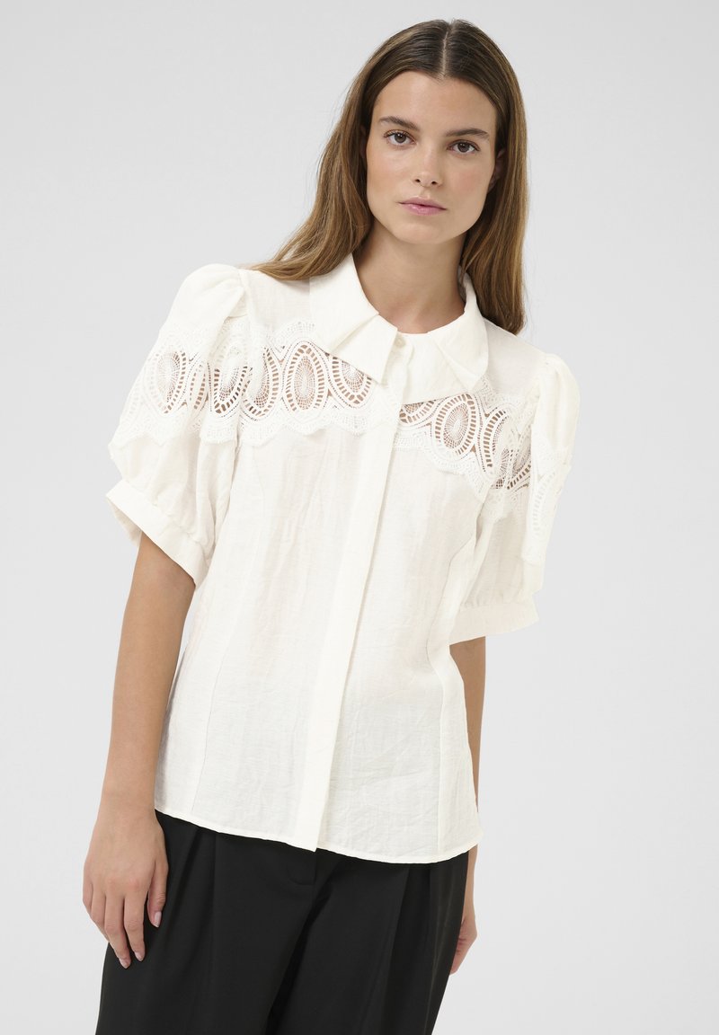 Blusa bianca in lino con maniche a sbuffo e chiusura a bottoni frontale, con dettagli di ricami in pizzo sulla parte anteriore. Design semplice e testurizzato.