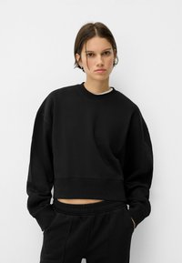 Črn skrajšan pulover z padlimi rameni, ribanim ovratnikom in mehko teksturo. Usklajen z vojaško črnimi hlačami, ki imajo elastičen pas.