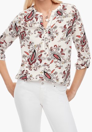 Nainen, jolla on valkoiset housut ja valkoinen pusero, jossa on punaisia, mustia ja oransseja kukallisia paisley-kuvioita, hihat käärittynä kyynärpäihin.