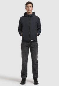 Marineblaue leichte Jacke mit Kapuze, Reißverschluss und zwei Seitentaschen. Kombiniert mit grauen Jeans und schwarzen Schuhen, stehend vor einem neutralen Hintergrund.