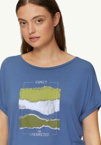 Niebieska koszulka z krótkim rękawem o luźnym kroju. Posiada graficzny nadruk zielonych i białych pasków z napisem "OCZEKUJ NIEOCZEKIWANEGO."