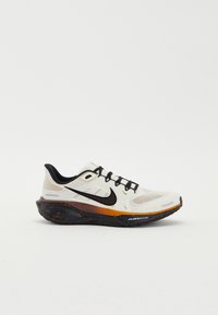 Chaussure de course Nike blanche et noire avec un swoosh noir, des accents orange et une semelle noire tachetée sur un fond uni.