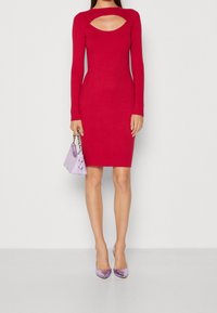 Robe en tricot côtelé rouge avec des manches longues et un détail découpé au niveau de la poitrine. Associée à un sac à main lavande et des talons violets assortis.