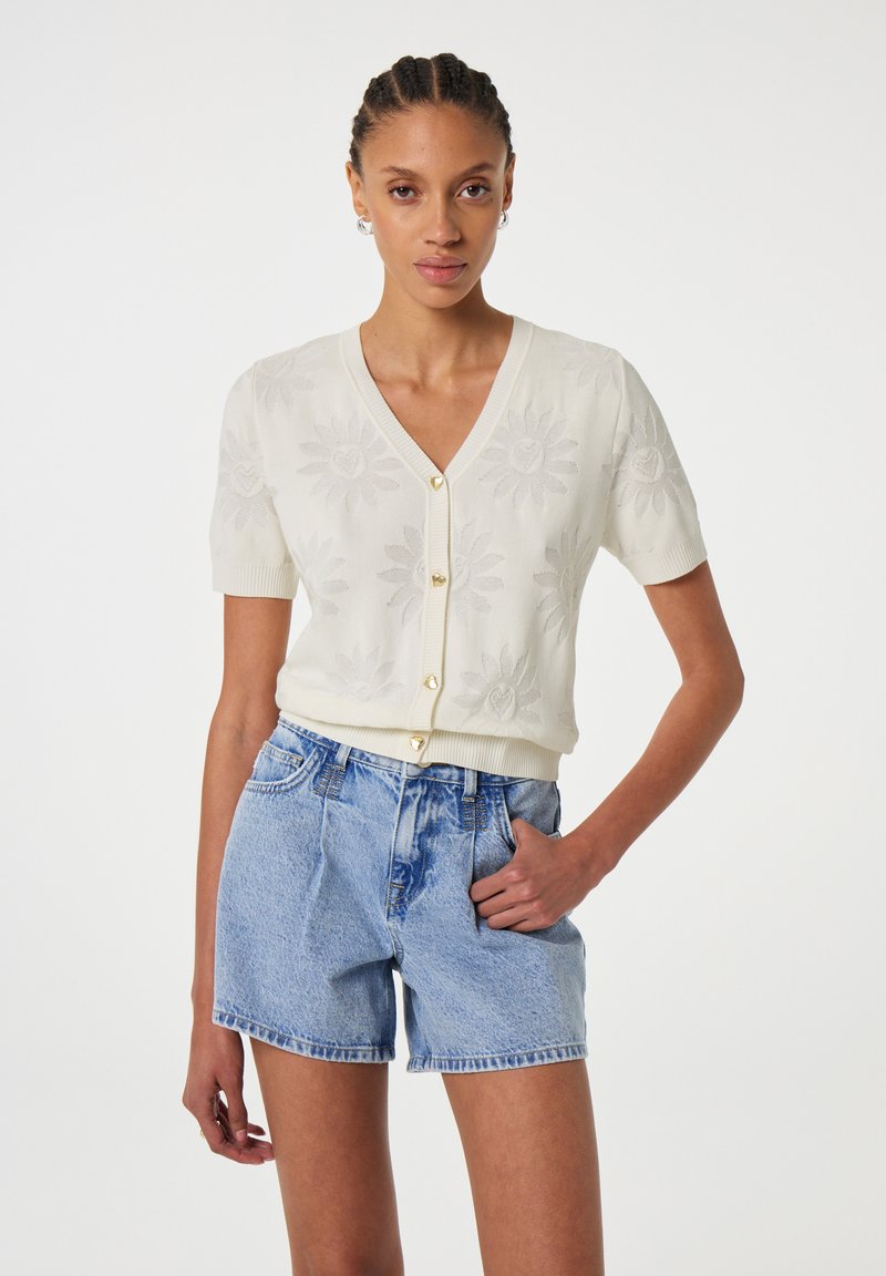 Crème cardigan met korte mouwen, voorzien van een gebloemd breipatroon en goudkleurige knopen, gecombineerd met lichtblauwe denim shorts.