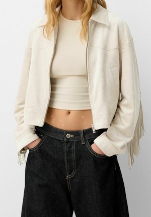 Young woman wearing a cropped beige jacket with fringe, beige crop top, and high-waisted black jeans with hands in pockets.