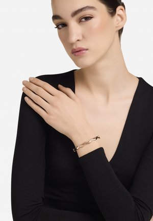 Rózsaarany csavart karkötő, amelyet egy modell csuklóján viselnek, fekete mély V-nyakú hosszú ujjú felsővel párosítva. Simított fémfelület, minimalista kialakítás.