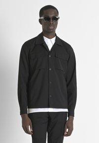 Giacca nera a bottoni con due tasche frontali, maniche lunghe e taglio dritto. Indossata sopra una camicia bianca, abbinata a occhiali da sole.