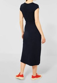 Granatowa sukienka do kolan z krótkimi rękawami, dopasowanym stanem i bocznym rozcięciem. Noszona z czerwonymi trampkami i beżową podeszwą w casualowym stylu.