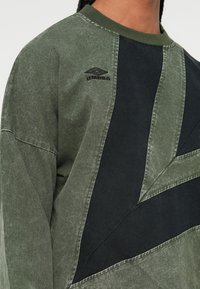 Zöld és fekete színblokkokkal díszített pulóvert viselő személy, akinek a mellkasán egy hímzett Umbro logó található.