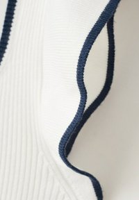 Tissu blanc côtelé avec un bord bleu marine formant un détail ondulé et volanté, probablement une partie d'une manche ou d'un col de vêtement.