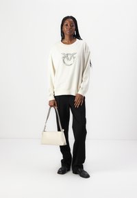 Krémová oversized mikina s logem z rhinestones, spárovaná s černými na míru šitými kalhotami. Modelka drží malou bílou kabelku s krátkým popruhem.