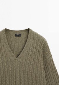 Olive green cable-knit sweater with a deep V-neck and ribbed cuffs, showcasing a textured pattern and a soft, knitted fabric.
