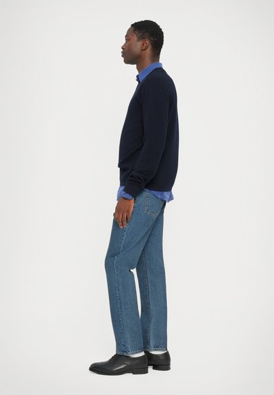 Marineblauwe trui over een lichtblauw shirt, gecombineerd met lichtblauwe jeans en zwarte schoenen. Stof lijkt zacht met een ontspannen pasvorm.