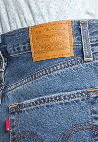 Jeans in denim con un'etichetta in pelle color tan con loghi impressi. Il tessuto blu mostra cuciture classiche e un'etichetta rossa sulla tasca posteriore.