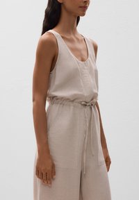 Beige ärmelloser Jumpsuit mit tiefem Ausschnitt, taillierter Taille, verstellbarem Band und zwei vorderen Taschen; aus einem leichten Stoff gefertigt.