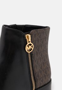 Gros plan sur une botte cheville noire et marron avec une fermeture éclair dorée et un tirant rond au logo MK.