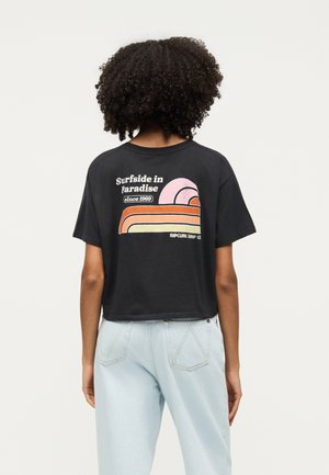 Άτομο με σγουρά μαλλιά που φοράει μαύρο κοντό μπλουζάκι με το κείμενο «Surfside in Paradise since 1969» και ρετρό γραφικό κύματος, σε συνδυασμό με ανοιχτόχρωμα μπλε τζιν.