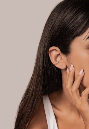 Gold hoop earring with a decorative floral accent, worn on the ear. Long, straight hair partially covers the earring, with a neutral background.