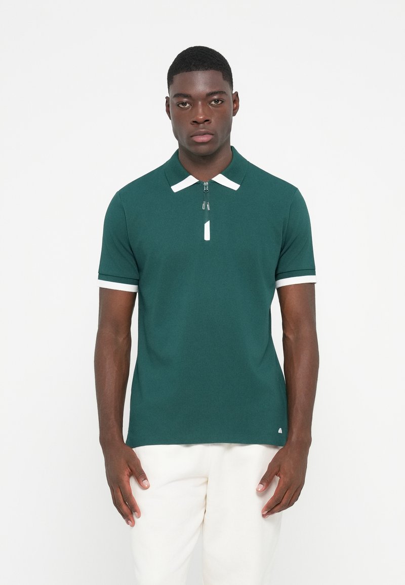 Grøn polo shirt lavet af et glat stof, med en lynlåse krave, hvide detaljer ved halsudskæringen og ærmerne, samt en tætsiddende pasform.